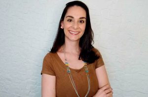 Mulher de cabelo escuro sorrindo, blusa marrom, em pose confiante, contra fundo claro.