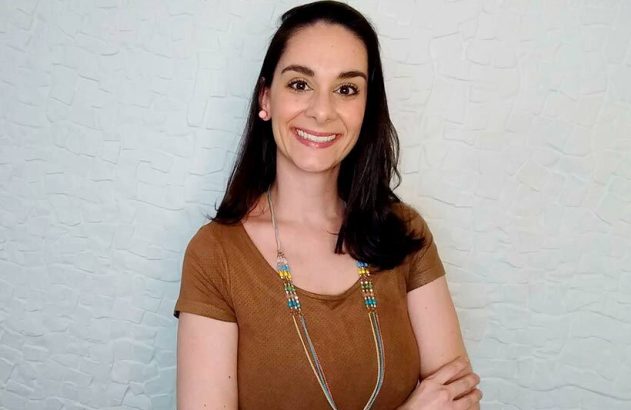 Mulher de cabelo escuro sorrindo, blusa marrom, em pose confiante, contra fundo claro.