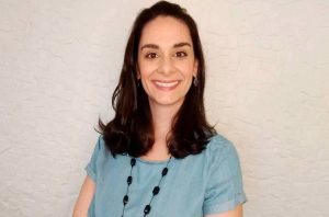 Mulher de cabelo escuro e blusa azul sorrindo para a câmera. Fundo branco.