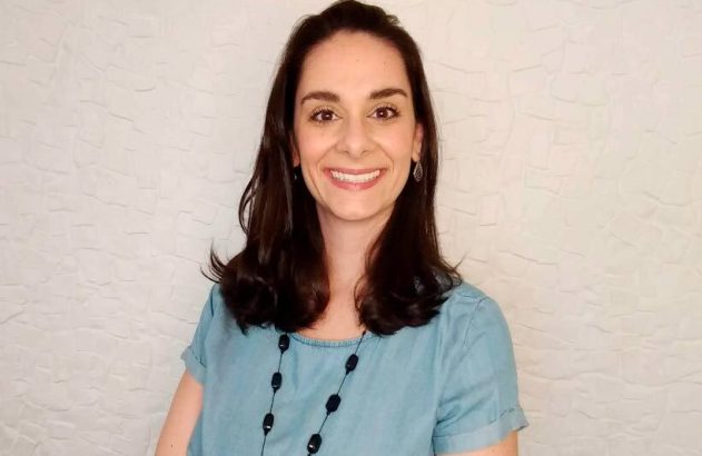 Mulher de cabelo escuro e blusa azul sorrindo para a câmera. Fundo branco.