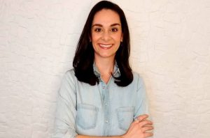 Mulher morena sorrindo, camisa jeans clara e braços cruzados.