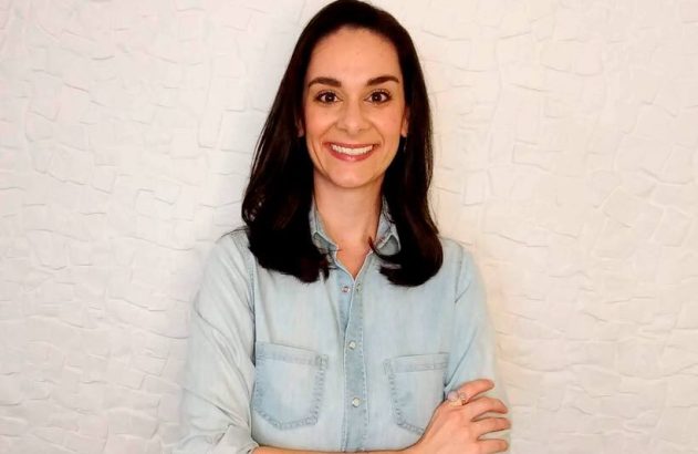 Mulher morena sorrindo, camisa jeans clara e braços cruzados.