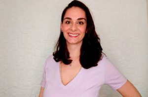 Mulher sorridente, cabelo escuro e blusa V-neck clara, em pé. Fundo neutro.