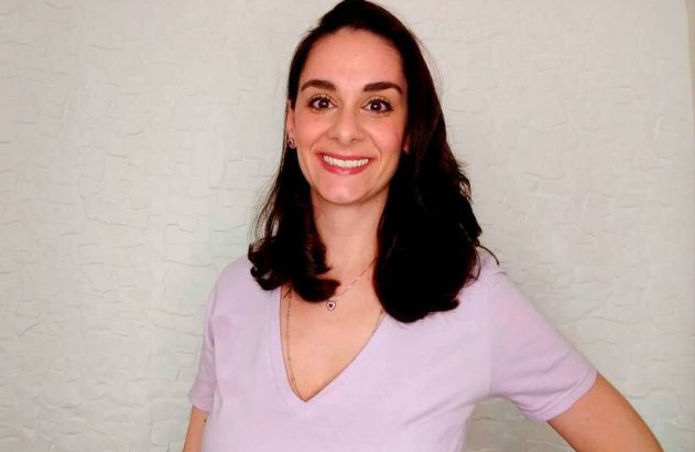 Mulher sorridente, cabelo escuro e blusa V-neck clara, em pé. Fundo neutro.