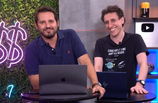 Dois homens sorrindo atrás de mesas com laptops em um estúdio com fundo verde e neon.