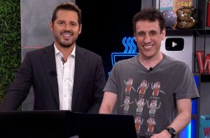Dois homens sorrindo para a câmera. Um veste terno, o outro camiseta estampada, em estúdio.