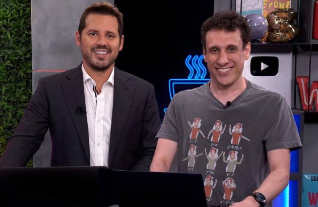 Dois homens sorrindo para a câmera. Um veste terno, o outro camiseta estampada, em estúdio.