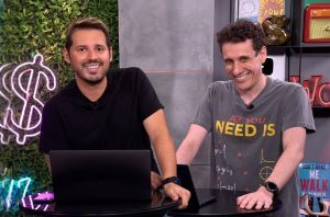 Dois homens sorrindo em uma mesa com laptops. Um de camisa preta, outro com camiseta "All You Need Is".