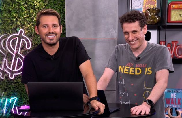Dois homens sorrindo em uma mesa com laptops. Um de camisa preta, outro com camiseta "All You Need Is".