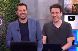 Dois homens sorridentes sentados em frente a laptops em um estúdio.