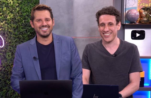 Dois homens sorridentes sentados em frente a laptops em um estúdio.