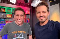Dois homens sorrindo posam para uma selfie em um estúdio com prateleiras e itens decorativos ao fundo.