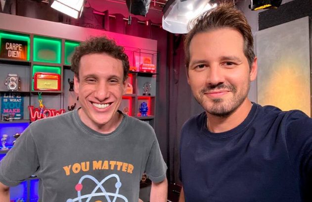 Dois homens sorrindo posam para uma selfie em um estúdio com prateleiras e itens decorativos ao fundo.