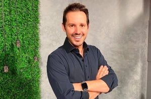 Homem sorridente com braços cruzados e camisa escura, olhando para a câmera.