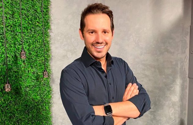 Homem sorridente com braços cruzados e camisa escura, olhando para a câmera.