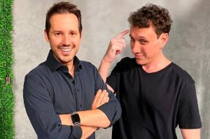 Dois homens. Um sorri com braços cruzados. O outro aponta para a própria cabeça em gesto pensativo.