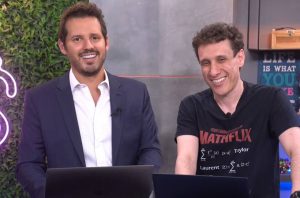 Dois homens sorrindo para a câmera, sentados em mesas com laptops. Um de terno, o outro de camiseta.