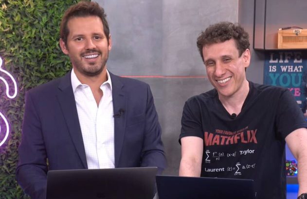 Dois homens sorrindo para a câmera, sentados em mesas com laptops. Um de terno, o outro de camiseta.