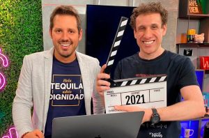 Dois homens sorridentes posam em estúdio. Um segura claquete de cinema exibindo '2021'.