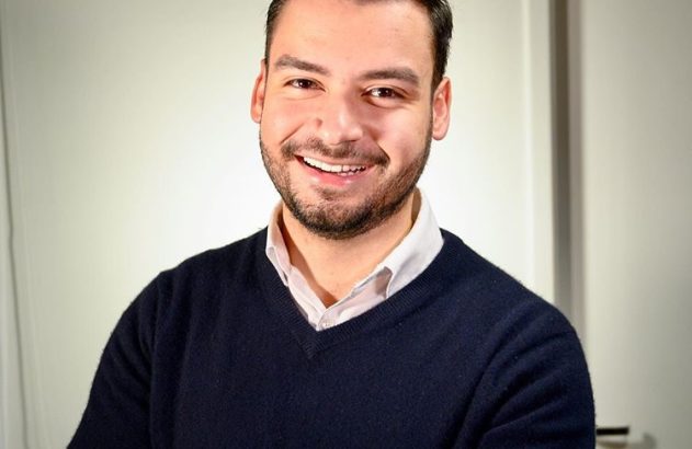Homem com barba sorrindo, de suéter escuro e camisa, braços cruzados. Parede clara ao fundo.