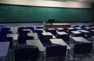 Sala de aula vazia com lousa verde cheia de anotações de economia e carteiras escolares azuis.
