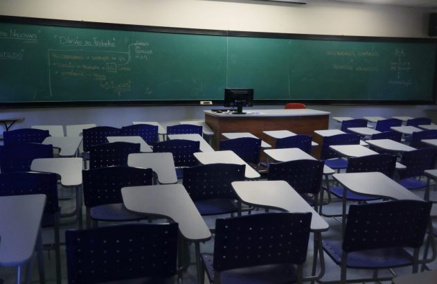 Sala de aula vazia com lousa verde cheia de anotações de economia e carteiras escolares azuis.