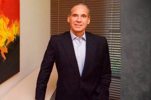 Homem de terno escuro, sorrindo, em pé, em ambiente profissional com persianas e pintura.