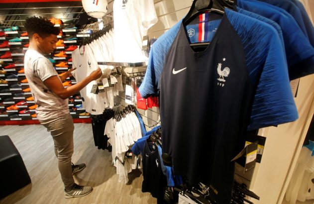 Jovem em loja de esportes examina camisa de futebol branca. Em primeiro plano, camisas azuis da seleção francesa com o galo e Nike.