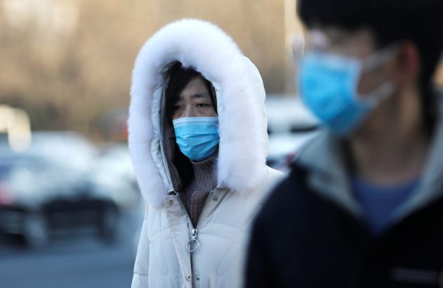 Mulher de casaco branco com capuz de pele e máscara azul. Outra pessoa mascarada desfocada à direita.