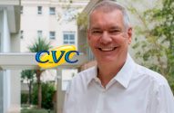 Homem sorridente de camisa branca, com logo CVC, em ambiente com arquitetura moderna e plantas.