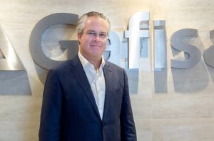 Executivo de terno e óculos sorri em frente a letras metálicas formando 'Gafisa' em uma parede clara.