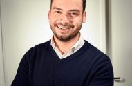 Homem com barba sorrindo e braços cruzados, vestindo suéter azul marinho e camisa clara.