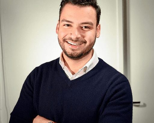 Homem com barba sorrindo e braços cruzados, vestindo suéter azul marinho e camisa clara.