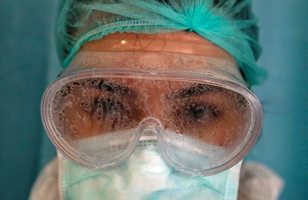 Close-up: Olhos de pessoa sob óculos de segurança embaçados pelo vapor, com touca e máscara médica.