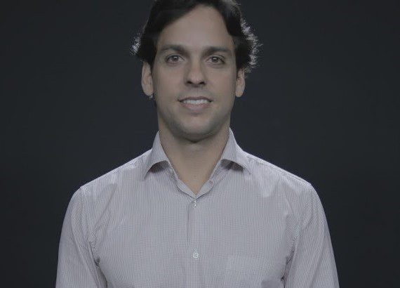 Retrato de um homem sorrindo com cabelo escuro e camisa clara contra um fundo escuro.