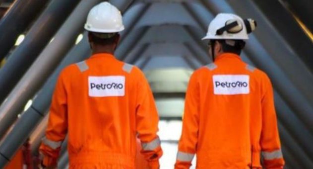 Dois funcionários da PetroRio, de costas, com macacões laranja e capacetes brancos, caminhando em área industrial.