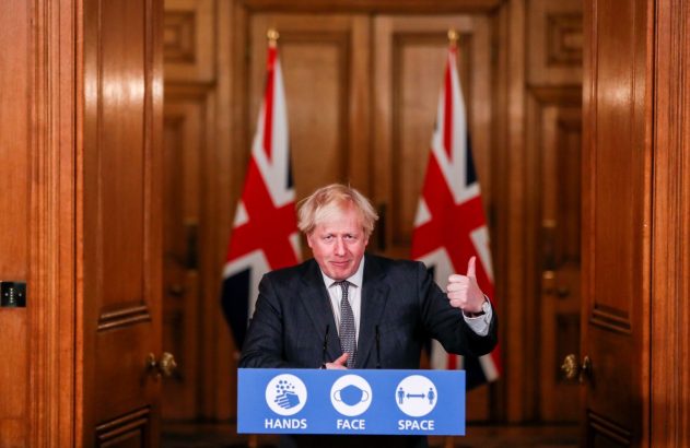 Boris Johnson, em pódio com bandeiras do Reino Unido e dicas "Mãos, Rosto, Espaço", faz um sinal de positivo.