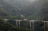 Caminhões trafegam em longo viaduto sobre mata atlântica exuberante e montanhas com cachoeira ao fundo.