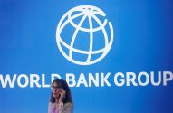 Painel azul com logo e nome "WORLD BANK GROUP" em branco. Mulher desfocada no primeiro plano falando ao celular.
