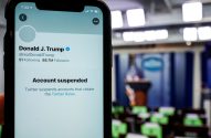 Smartphone exibe perfil de Donald Trump no Twitter com 'Conta suspensa'. Fundo borrado de sala de imprensa.
