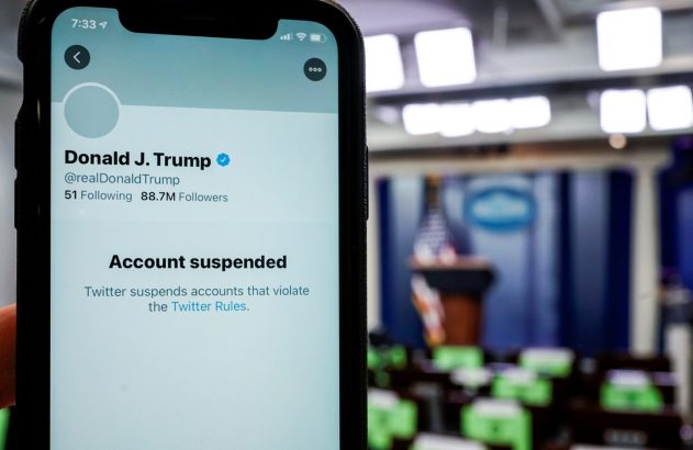 Smartphone exibe perfil de Donald Trump no Twitter com 'Conta suspensa'. Fundo borrado de sala de imprensa.
