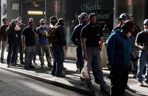 Homens com capacetes e cobrindo o rosto estão parados na rua. A luz do sol projeta sombras longas.