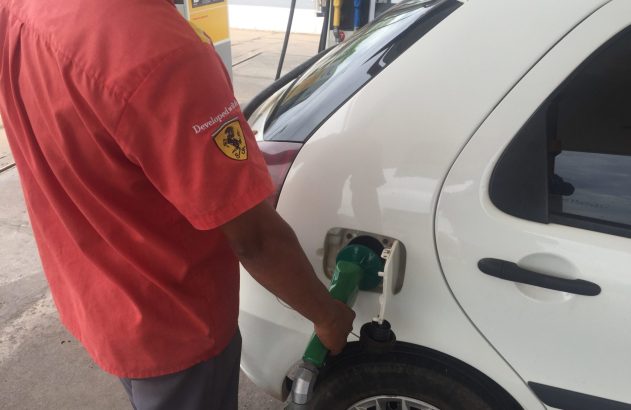 Frentista de camisa vermelha com logo Shell/Ferrari abastece carro branco em posto.