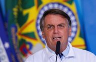 Jair Bolsonaro, em camisa branca, fala em microfone, com bandeira do Brasil desfocada ao fundo.