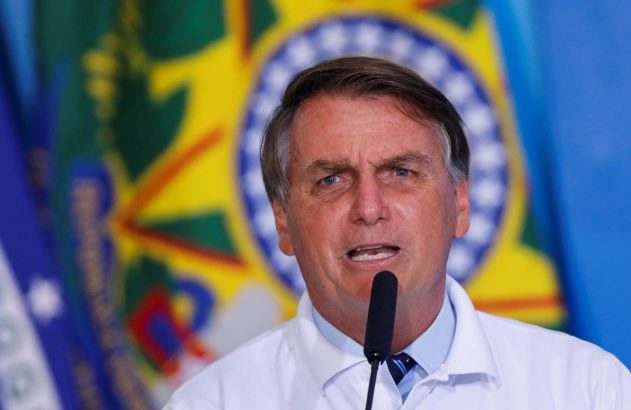 Jair Bolsonaro, em camisa branca, fala em microfone, com bandeira do Brasil desfocada ao fundo.