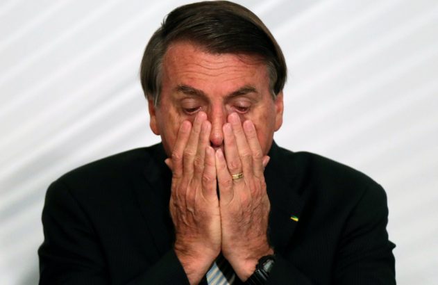 Homem de terno com as mãos sobre o rosto, olhos avermelhados, aparentando choro ou profunda tristeza.