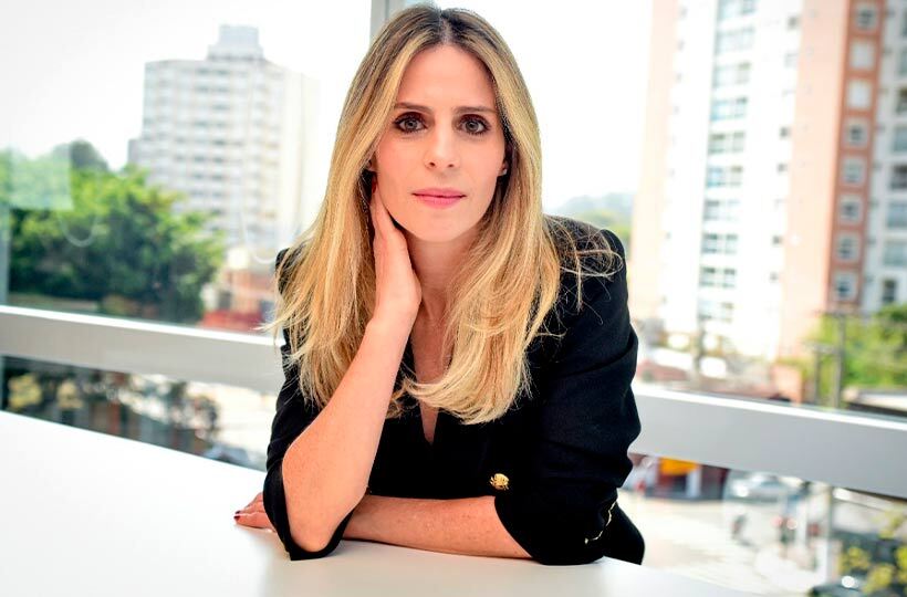 Mulher loira de blazer preto, braços na mesa e mão no rosto, olhando para frente. Fundo urbano.