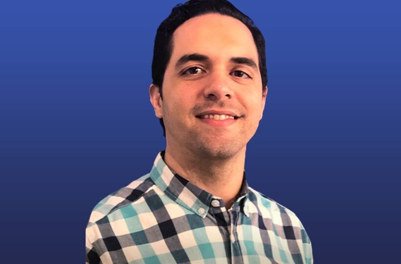Homem sorrindo com camisa xadrez azul e branca, contra um fundo azul.