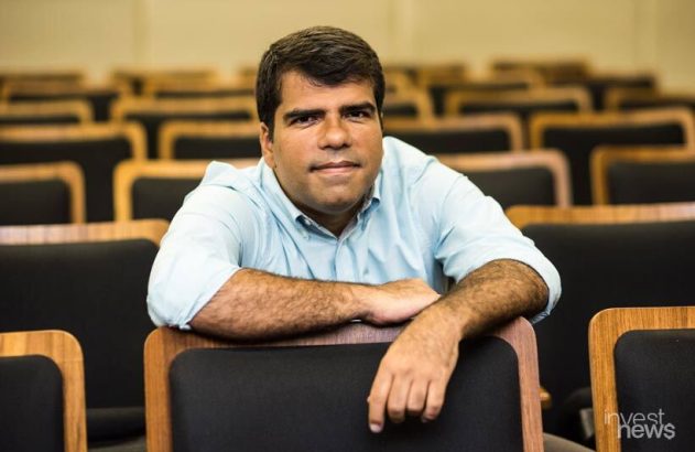 Homem de camisa clara, apoiado em cadeira de auditório, olhando para a câmera.