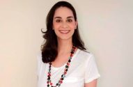 Mulher de cabelo escuro, camiseta branca e colar colorido sorri.